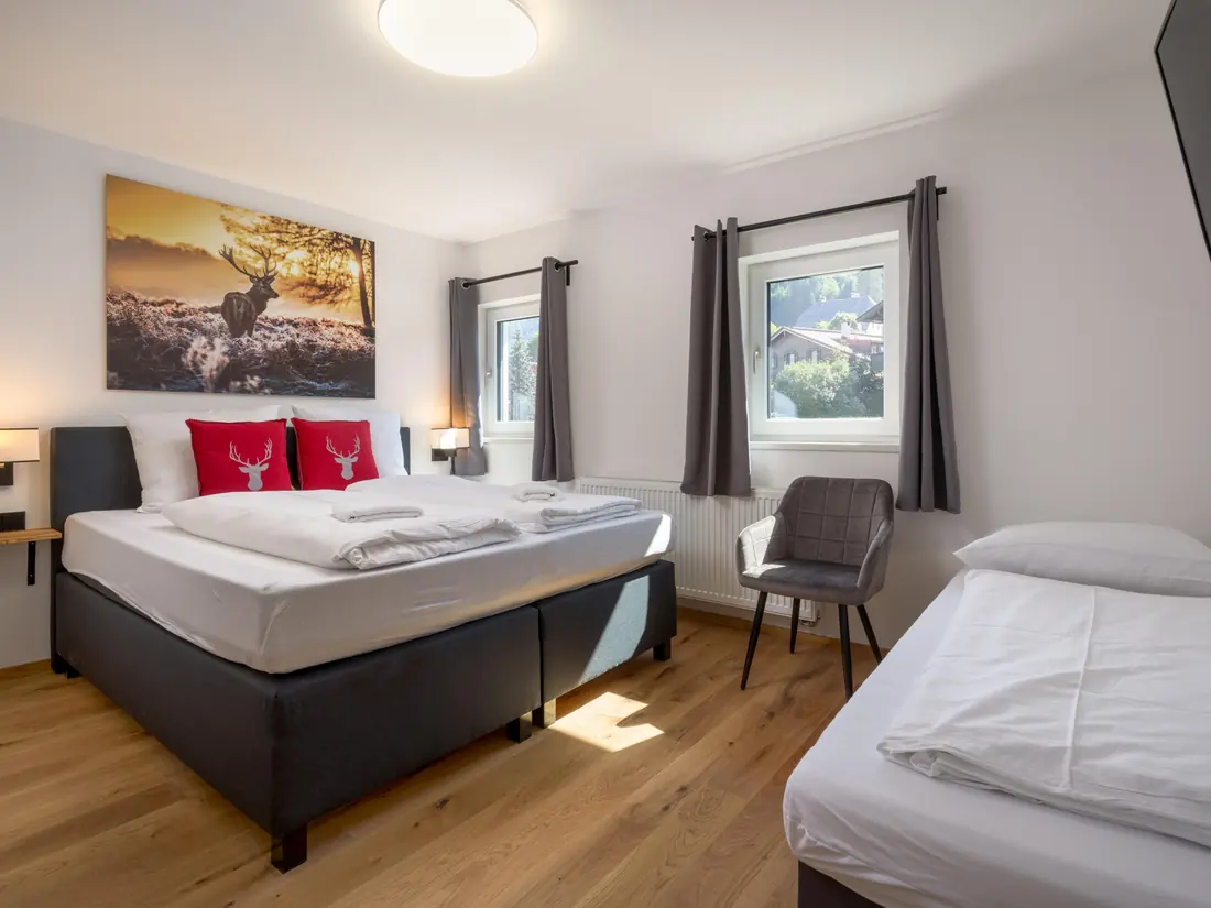 Een slaapkamer met een tweepersoonsbed, rode kussens met een hert, een eenpersoonsbed en twee ramen.