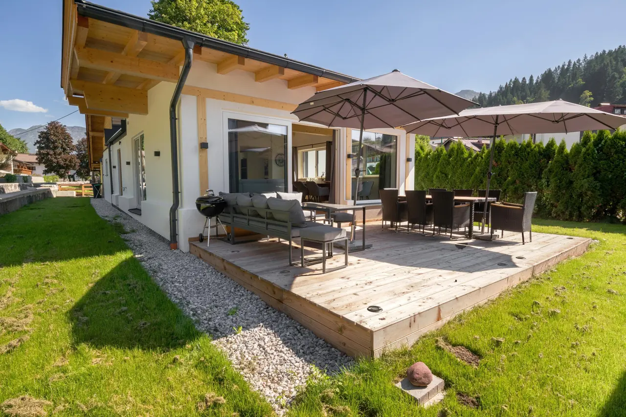 Het houten terras in de zomer met twee parasols, een eettafel, loungebank en houtskoolbarbecue, met gras eromheen.